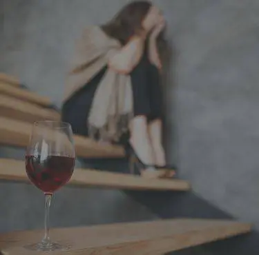 En person sitter hopkrupen i en trappa med händerna för ansiktet. I förgrunden syns ett glas vin.
